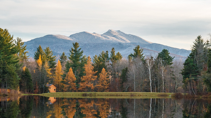 Landscape photo of a lake in the foreground surrounded by trees in fall with a mountain in the background.